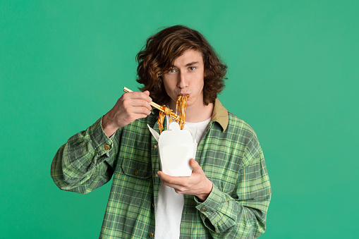 Food delivery concept. Young guy eating takeaway noodles on color background, blank space for design