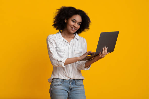istock Online Courses. Smiling Black Woman Using Laptop, Standing On Yellow Background 1211951350