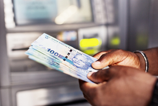 The hands of an African man hold a sheaf of South African One Hundred Rand banknotes next to an ATM.