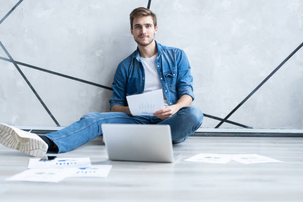 istock Man working at home. Handsome young man sitting on the floor and examining document while laptop and documents laying near him. 1211953079