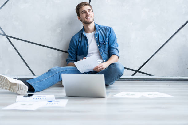 istock Man working at home. Handsome young man sitting on the floor and examining document while laptop and documents laying near him. 1211953080