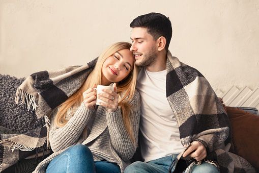 Happy young guy hugging his girlfriend under warm plaid at home