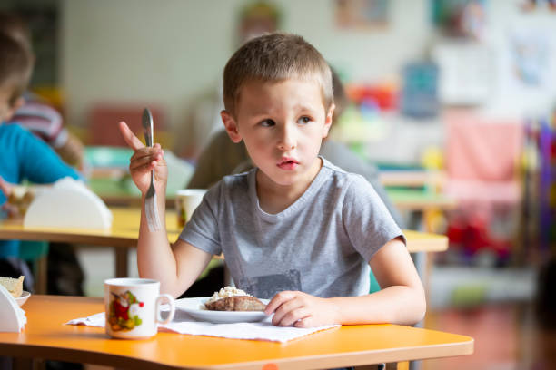 uma criança come no jardim de infância. um garotinho com pouco apetite. café da manhã para crianças. criança com um prato e garfo. - little boys breakfast caucasian child - fotografias e filmes do acervo