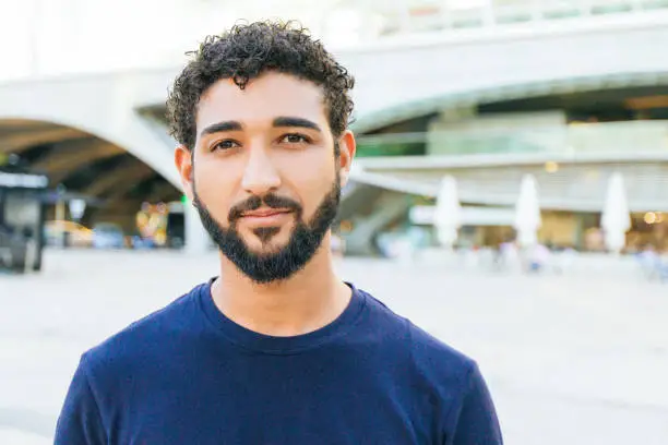 Pensive positive mix raced guy posing in city square. Front portrait on young man with black curly hair and beard wearing blue casual shirt, looking and smiling at camera. Male portrait concept