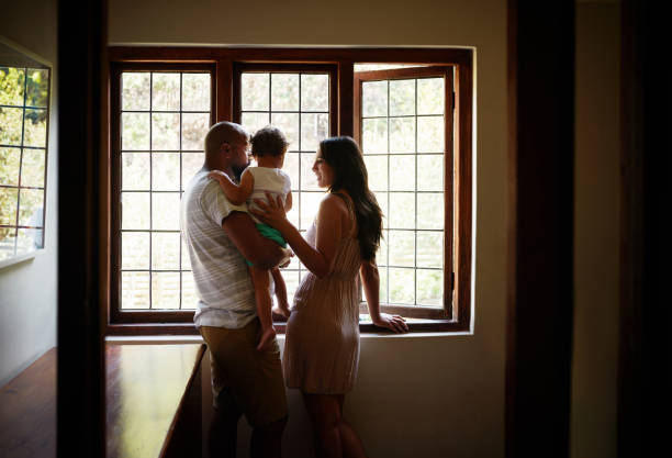 Nothing is more comforting than home and family Shot of a young family looking out of the window in their new home young family stock pictures, royalty-free photos & images
