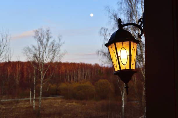 Lighted outdoor lamp against the beautiful spring landscape with the moon in sky and birch grove during sunset. Russia, Moscow region. Focus on foreground