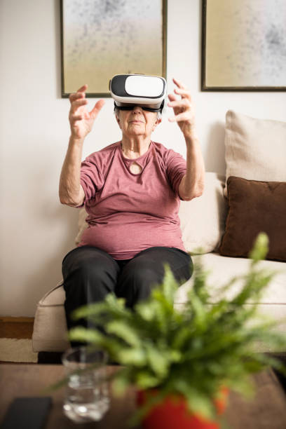 istock Senior woman sitting on a sofa in her living room and using virtual reality simulator goggles 1211965812