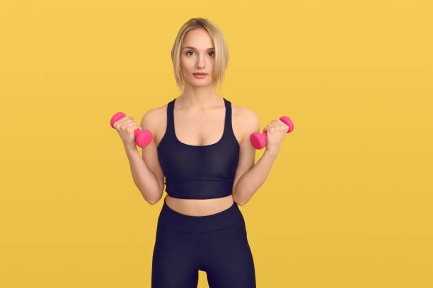 Pretty blond woman in black training top with small pink dumbbells in her hands, looking at camera. Close-up front portrait against plain yellow background