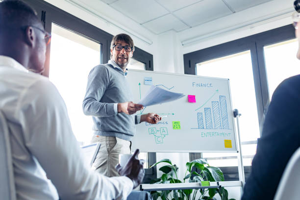 istock Handsome businessman pointing to whiteboard while explaining to the project to his colleagues on coworking place. 1211967982