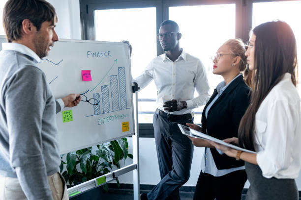 istock Handsome businessman pointing to whiteboard while explaining to the project to his colleagues on coworking place. 1211968039
