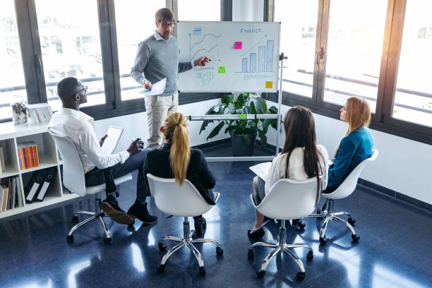 istock Handsome businessman pointing to whiteboard while explaining to the project to his colleagues on coworking place. 1211968070