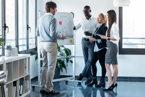 istock Handsome businessman pointing to whiteboard while explaining to the project to his colleagues on coworking place. 1211968098