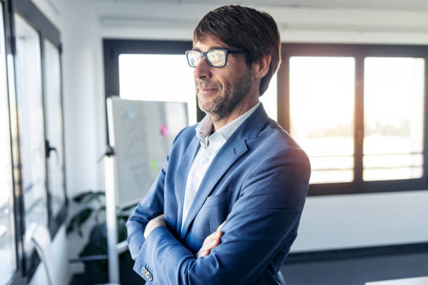 istock Handsome businessman with eyeglasses looking through the window while standing in the office. 1211968166