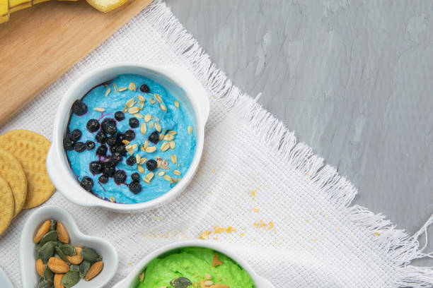 Two curd desserts: one with blueberries and nuts and the other with avocado and kiwi, next to there are pumpkin seeds and nuts