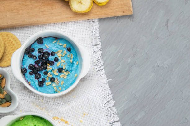 Two curd desserts: one with blueberries and nuts and the other with avocado and kiwi, next to there are pumpkin seeds and nuts