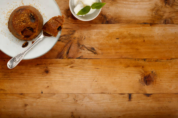 An individual spotted dick sponge cake, on a white plate with a spoon, and a small bowl of ice cream, on a wooden surface with copy space