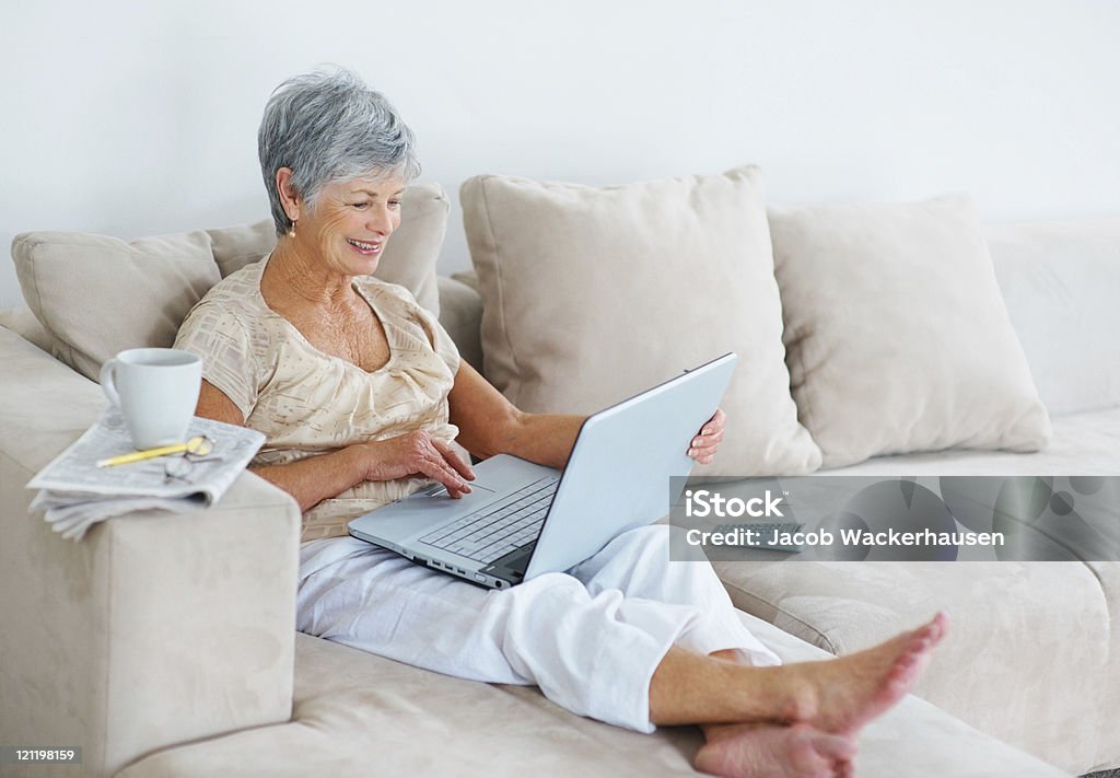 Улыбается Старший женщина, используя ноутбук, сидя на диване - Стоковые фото Газета роялти-фри