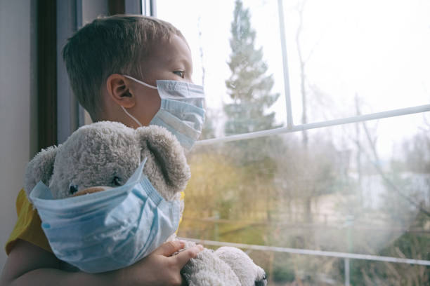 Sad illness child on home quarantine. Boy and his teddy bear both in protective medical masks sits on windowsill and looks out window. Virus protection, coronavirus pandemic, prevention epidemic. Sad illness child on home quarantine. Boy and his teddy bear both in protective medical masks sits on windowsill and looks out window. Virus protection, coronavirus pandemic, prevention epidemic. pandemic illness stock pictures, royalty-free photos & images