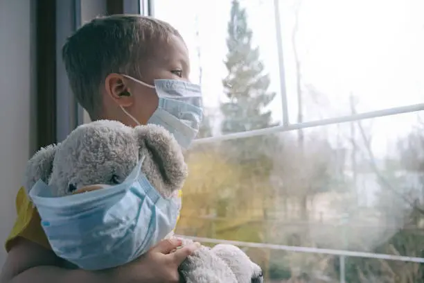 Photo of Sad illness child on home quarantine. Boy and his teddy bear both in protective medical masks sits on windowsill and looks out window. Virus protection, coronavirus pandemic, prevention epidemic.