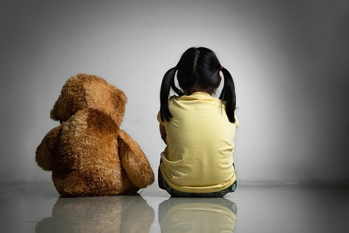 Little girl with teddy bear sitting on floor at empty room. Mental and depressed family concept. Back view
