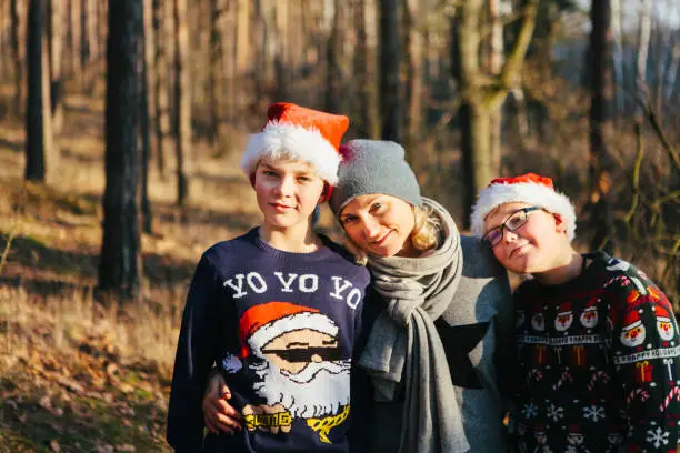December 1, 2018 - Kampinos, Poland:Pretty blond woman in warm clothing hugging close her two sons - teenage boys in Christmas sweaters and Santa hats, being silly, funny, together, standing in a sunny sunlit pine forest at sunset