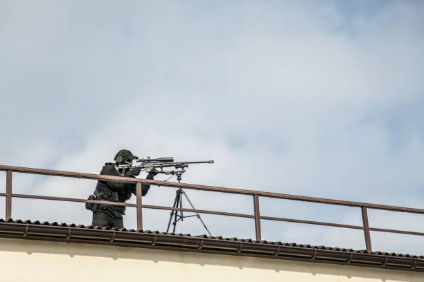 Police sniper aiming with optical sight from roof Police special operations or security team sniper standing on roof of city building and aiming with telescopic optical sight on sniper rifle mounted on tripod. SWAT shooter engage long range targets sniper stock pictures, royalty-free photos & images