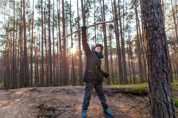 December 1, 2018 - Kampinos, Poland: winter fairy tale - a beautiful boy backlit in pine trees forest lifting his stick - imaginary weapon in a sign of victory