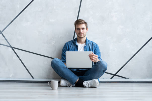 istock Young man working on laptop sitting on floor. Guy with computer reading news, surfing net or freelancing. 1212005434