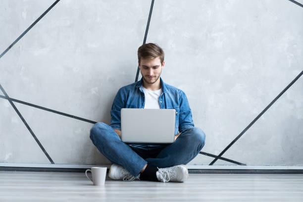 istock Young man working on laptop sitting on floor. Guy with computer reading news, surfing net or freelancing. 1212005467