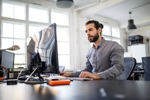 Young businessman sitting at his work desk and working on desktop computer. Male executive looking at camera and smiling while working on computer..