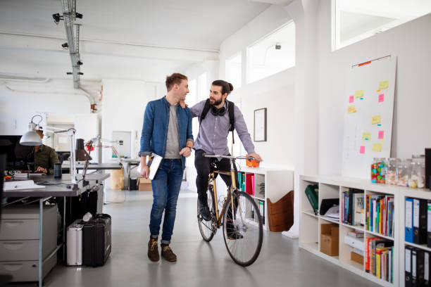 Office workers discussing work on the move Mid adult businessman walking with a colleague riding a bicycle in office. Two office workers on the move in office corridor and discussing work. hipster bike stock pictures, royalty-free photos & images
