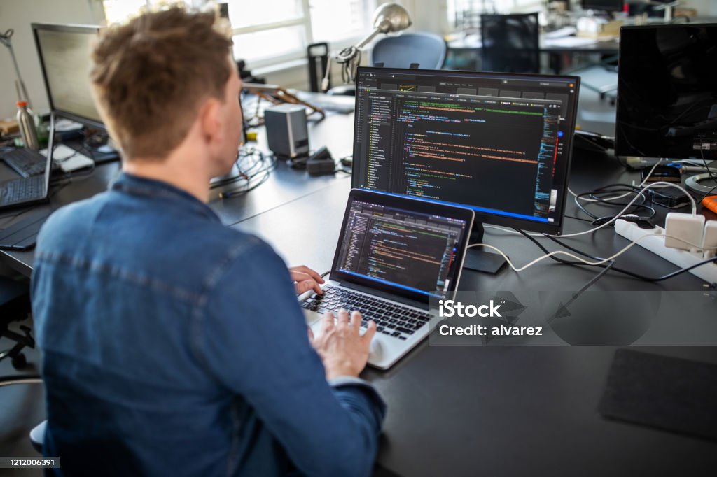 Computer programmer working on new software program Businessman working on laptop and looking at desktop computer monitor. Male professional sitting at his office desk and working on new software program. Technology Stock Photo