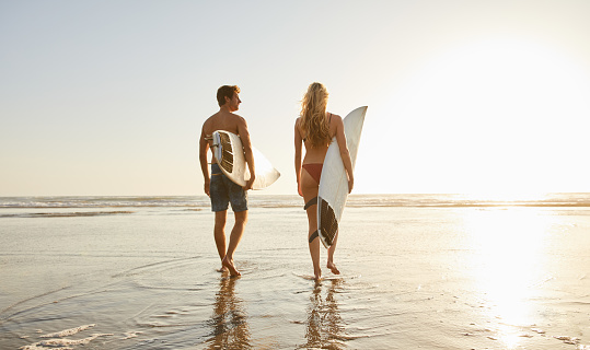 Young couple in swimwear carrying surfboards while walking on a sandy beach toward the ocean on a sunny day