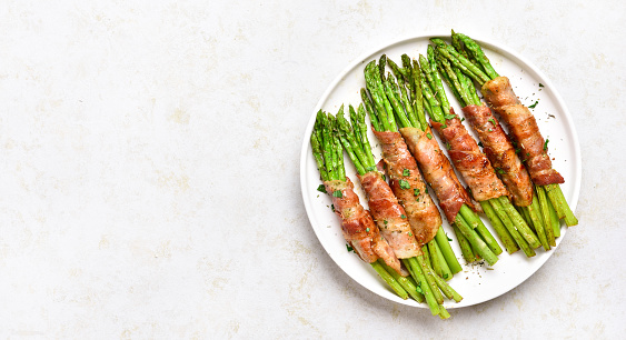 Bacon wrapped asparagus on white plate over light stone background with free text space. Top view, flat lay