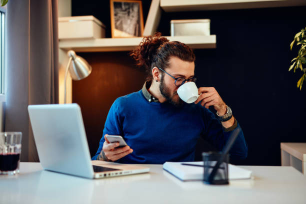 istock Young handsome bearded hipster sitting at his home office, holding cup of coffee and using smart phone. 1212009198