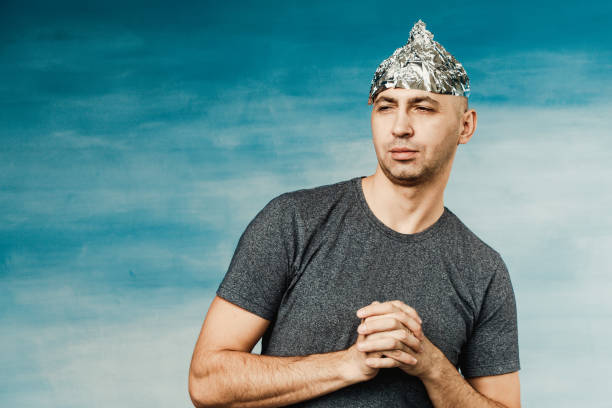 a man in a foil hat winks suspiciously and looks away on a blue background - tin foil hat imagens e fotografias de stock