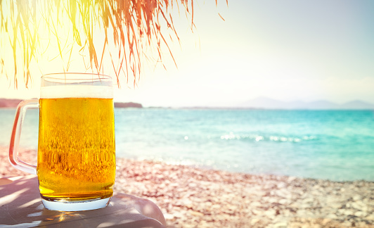 Cold glass mug of beer on beach on table under palm tree parasol