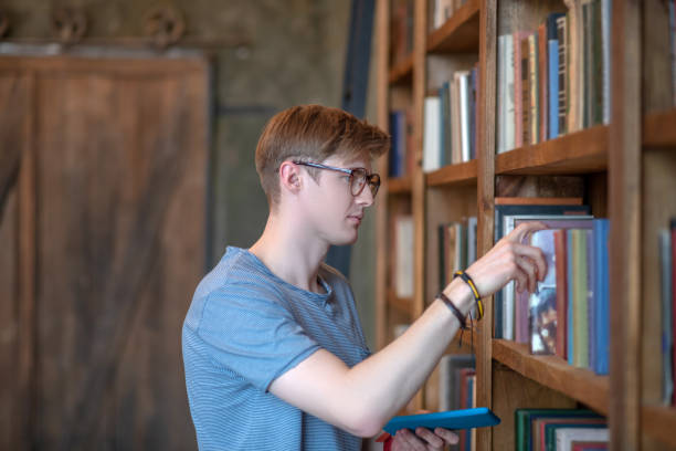 istock Young man in eyewear taking a book from a book shelf 1212035730