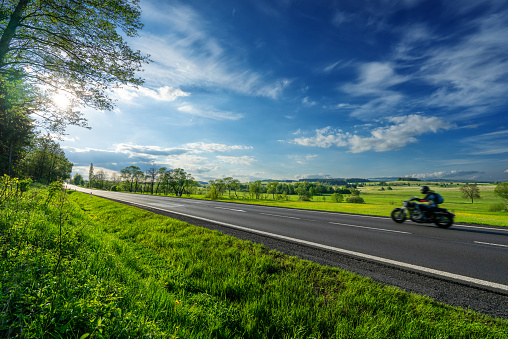 Black motorcycle riding on an empty asphalt road in a rural landscape under a radiant sun and dramatic clouds