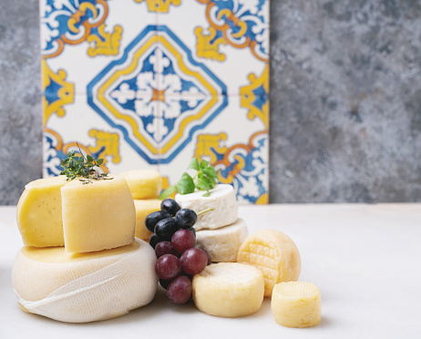 Traditional Portuguese semi-soft cheeses from evora alentejo and azores regions on the tray, served with fresh grapes, honey and herbs. Side view. Selective focus. Azulejo pattern.