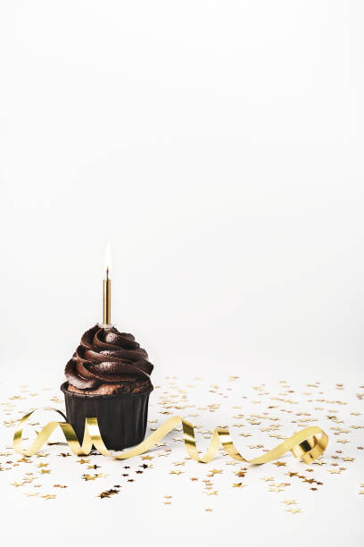 istock Chocolate cupcake with chocolate icing, sprinkled gold sparkles and burning candles on white background with golden star shaped confetti and party streamer. Holiday concept. Mockup for your design. Close-up. Copy space. 1212049694