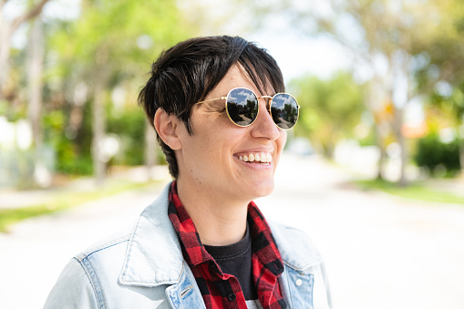 This is a photograph of a Caucasian gender neutral person with short hair in their 30s standing outside wearing a denim jacket and sunglasses on a sunny spring day in Miami, Florida, USA.