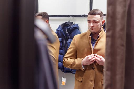 30 years old man trying on an autumn brown classic coat in a fitting room, collection sale