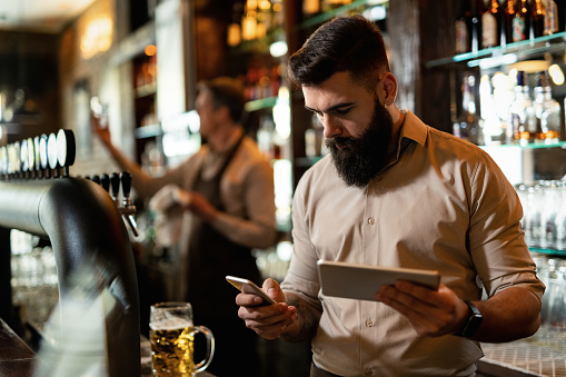 Young barista texting on smart phone while using touchpad in a bar.