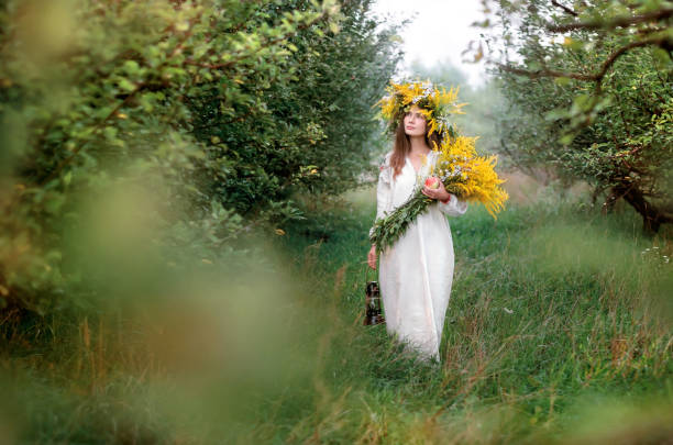 Young beautiful woman in a wreath of wildflowers and a long white national shirt Young beautiful woman in a wreath of wildflowers and a long white national shirt walks in the apple orchard. Ukrainian culture, ethno style slavic culture stock pictures, royalty-free photos & images