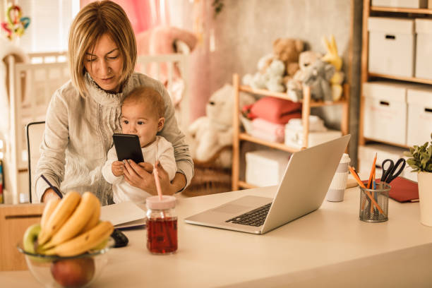 istock Woman holding her baby daughter and using her smart phone while working 1212076479