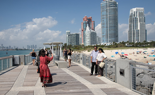 A middle aged asian couple pose for their picture being taken by mature asian woman as her companion looks on and a young caucasian couple approaches from their right.  This takes place on Miami’s south pointe park pier on an early march afternoon against a backdrop of high rise luxury condos with assorted beachgoers laying in the sand below.  The south miami skyline is in the far background