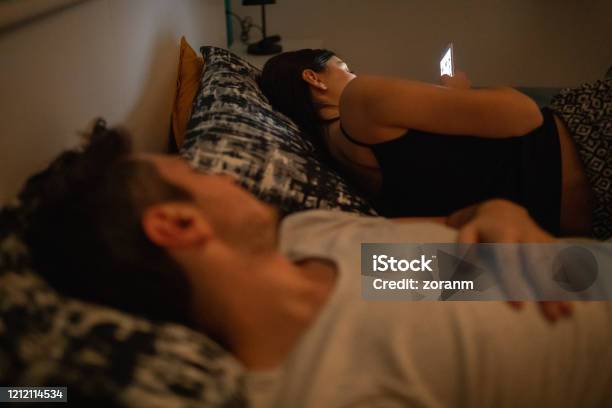 Young Woman Using Phone In Bed While Her Husband Is Asleep Stock Photo - Download Image Now