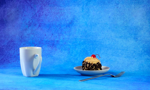 A high white mug with tea, next to a saucer with a biscuit cake in chocolate chips and cherry on a cream and a teaspoon.