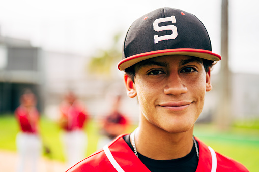 Portrait of relaxed young Hispanic baseball player standing outdoors on the field with teammates and waiting for game time.